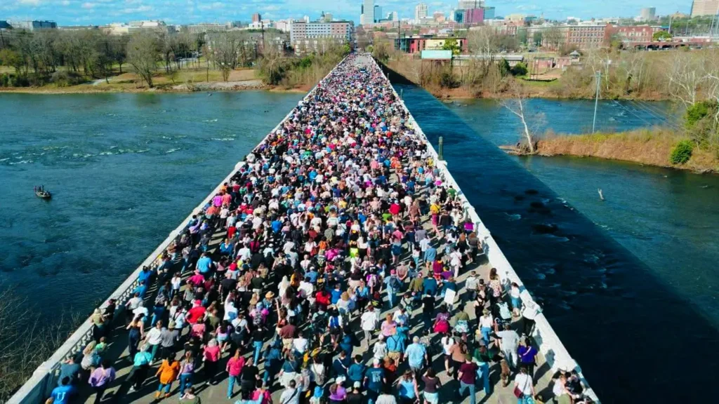 “Large crowd standing on a bridge observing Buddhist monks during the Walk for Peace, symbolizing public support, harmony, and peaceful unity.”